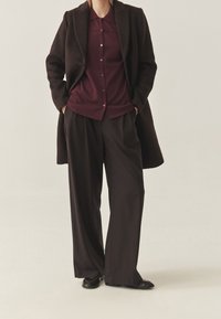 Chemise boutonnée couleur bordeaux avec col, portée sous un manteau marron foncé. Pantalon ample de couleur foncée, associé à des chaussures noires.