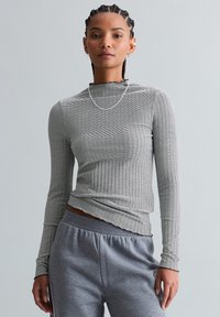 Vrouw met ingevlochten haar, die een zwart-wit gestreept shirt met lange mouwen en grijze joggingbroek draagt, staand tegen een effen grijze achtergrond.