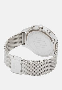 Montre en acier inoxydable argenté avec un bracelet en maille, un boîtier rond et un dos en verre mettant en valeur une étiquette gravée et deux boutons latéraux.