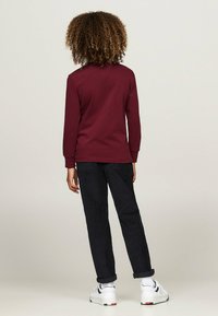 Felpa color bordeaux con maniche lunghe, abbinata a pantaloni in velluto a coste neri. Sneakers bianche con dettagli neri e calzini alla caviglia. Sfondo semplice.
