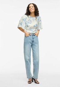 Blusa azzurra con motivo floreale e maniche a sbuffo, abbinata a jeans in denim chiaro a vita alta e gamba dritta, e sandali neri con tacco.