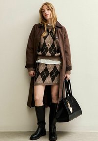 Femme portant un pull en argyle marron et une jupe, accompagnée d'un long manteau marron, de bottes noires jusqu'aux genoux, d'un collier, et tenant un grand sac à main noir.