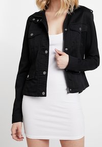 Žena v přiléhavých bílých šatech bez rukávů s černou bundou na knoflíky a se zipem přes ně, stojící proti jednoduchému pozadí.