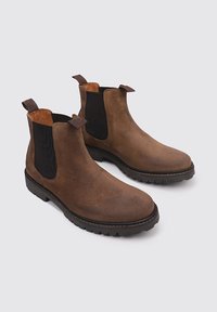 Botas Chelsea de ante marrón con paneles elásticos laterales negros, puntera redonda, lengüetas para tirarlas y suelas de goma resistentes. Sutiles acentos de costura.