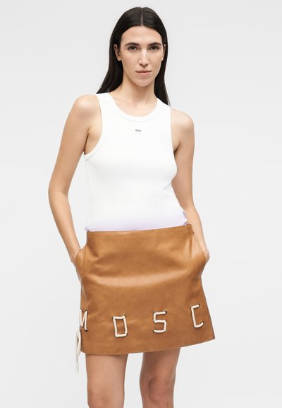Γυναίκα που φοράει ένα λευκό αμάνικο τοπ και καφέ δερμάτινη φούστα με τα γράμματα "MOSC" από σκοινί μπροστά, χέρια στις τσέπες.