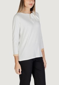 Maglietta bianca a maniche lunghe con dettaglio a fiocco sullo scollo e un accento di tessuto a contrasto nei polsini. Tessuto morbido, vestibilità rilassata. Pantaloni neri.