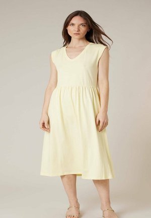 Femme debout portant une robe sans manches jaune pâle, longueur genou, avec un col en V, et des sandales beige à lanières, sur un fond uni.