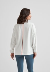 Woman standing turned away wearing a white long-sleeve top with central vertical brown stripe and blue fitted pants.