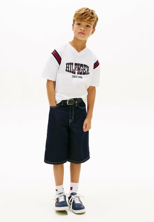 Niño con cabello rubio vistiendo una camiseta blanca de manga corta con rayas rojas y pantalones cortos azul oscuro hasta la rodilla, de pie sobre un fondo blanco.