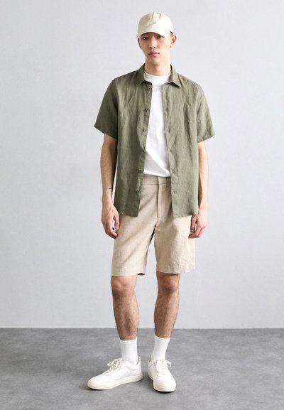 Chemise en lin vert olive à manches courtes, t-shirt blanc à col rond, shorts beiges, baskets blanches et casquette crème, se tenant devant un fond neutre.