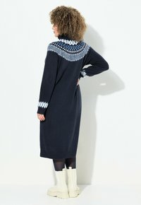 Donna con capelli ricci che indossa un vestito a maglia blu navy con yoke e polsini a fantasia bianca e blu, collant neri e stivali bianchi, in piedi di profilo.