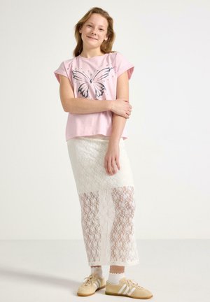 Pige med lys brunt hår iført en lyserød T-shirt med sommerfuglemotiv, hvid blondenederdel, hvide sokker og beige sneakers, står og smiler.