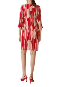 Figurbetontes Kleid mit rot-beigem abstraktem Muster, drei Viertel Ärmeln und Reißverschluss im Rücken. Kombiniert mit roten High Heels.