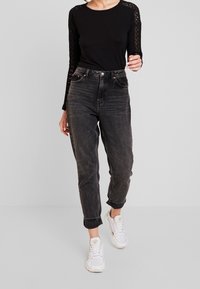 Schwarzes Langarmoberteil mit Spitzenverzierungen an den Ärmeln, kombiniert mit hochgeschnittenen, ausgewaschenen schwarzen Jeans und weißen Sneakers. Lässiges Outfit.