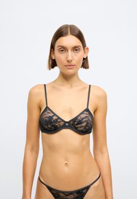 Crna čipkasta braletica s tankim trakicama, izrađena od prozirnog materijala i s suptilnim cvjetnim uzorkom. Središnji detalj s hardverom dodaje estetski naglasak dizajnu.