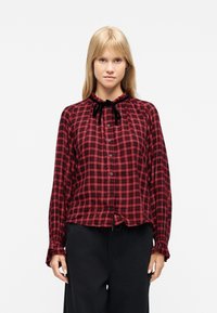 Νεαρή γυναίκα με ξανθά μαλλιά που φοράει κόκκινη και μαύρη καρό μπλούζα με κουμπιά και μαύρη κορδέλα στο γιακά, καθώς και μαύρο παντελόνι.