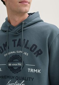 Hallhimu sinine kapuutsiga sviiter puuvillase seguga. Omab nööre, suurt graafilist trükist tumesinises ja valges ning pehmet tekstuuri.