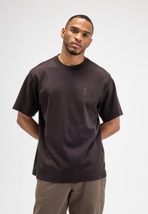 Mann mit kurzen Haaren und Bart, der ein dunkelbraunes kurzärmeliges T-Shirt und hellbraune Hosen trägt, steht vor einem schlichten hellen Hintergrund.