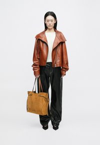 Jachetă din piele maro cu guler lat, pulover crem, pantaloni negri larghi și geantă din suede bej cu închidere tip șnur și bretele pentru umăr.