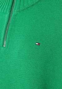 Groene zip-up trui met geribde kraag, gestructureerd breipatroon en een klein geborduurd logo met rood, wit en blauwe strepen.