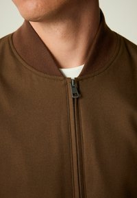 Braune Zip-Jacke mit geripptem Kragen und glatter Stofftextur. Der Reißverschluss ist silberfarben und befindet sich in der Mitte vorne.