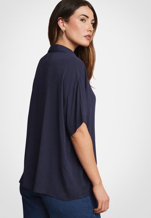 Donna con capelli castani lunghi che indossa una camicetta blu navy a maniche corte e larghe e jeans blu, vista di profilo su uno sfondo semplice.