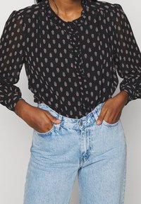 Sort bluse med små røde og hvide blomster, rynkede foran og lange ærmer, sammen med lyseblå high-waisted jeans.