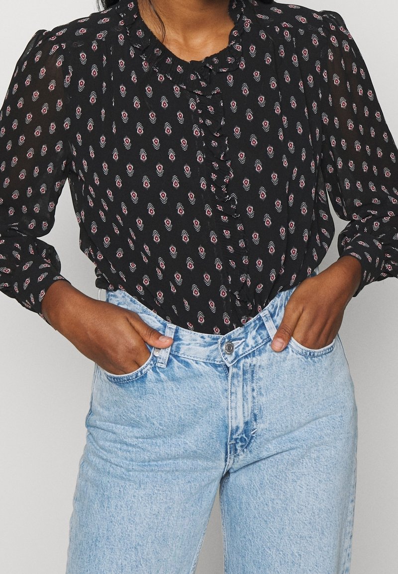 Sort bluse med små røde og hvide blomster, rynkede foran og lange ærmer, sammen med lyseblå high-waisted jeans.