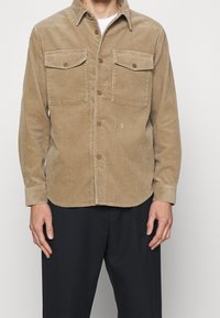 Camisa de pana en marrón claro con cierre de botones en el frente, dos bolsillos en el pecho y mangas largas. Presenta un cuello puntiagudo y una textura sutil.