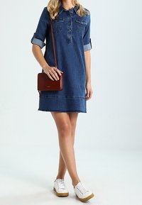 Vestido de mezclilla con cuello, mangas cortas enrolladas, dos bolsillos en el pecho, largo hasta el muslo, combinado con un bolso de bandolera rojo y zapatillas blancas.