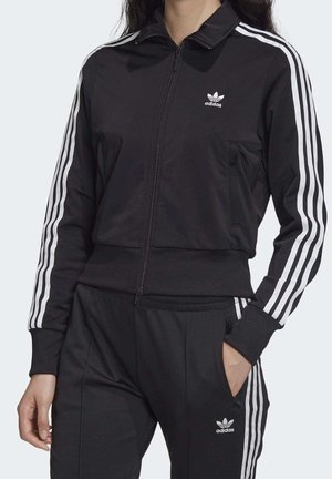 Ensemble de sport noir avec veste zippée, accents blancs à 3 bandes sur les manches et les pantalons, comprenant un logo brodé sur la poitrine et la cuisse.