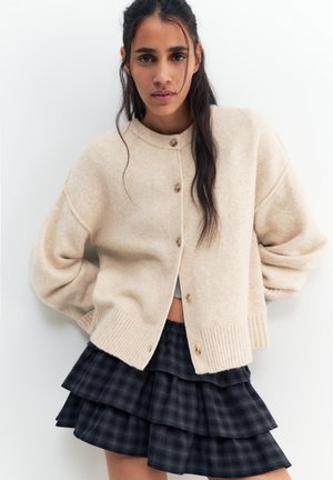Ung kvinde med langt mørkt hår iført en beige cardigan med knapper og et lagdelt mørkt ternet skørt, poserer mod en ensfarvet hvid baggrund.