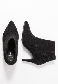 Botines negros con puntas afiladas, tacones altos, textura con tachuelas y cremallera lateral, mostrados desde vistas superior y lateral sobre un fondo blanco.