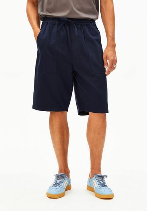 Mann trägt marineblaue Shorts mit Kordelzug und Vordertaschen sowie hellblaue Turnschuhe mit Gummisohlen, steht vor weißem Hintergrund.