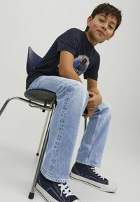 T-shirt graphique bleu avec un design sur le devant, jeans droit bleu clair, et baskets navy avec des lacets blancs, assis sur une chaise.