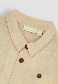 Camisa de lino beige con cuello, botones marrones y un bolsillo en el pecho. La etiqueta indica "Jo-Jo Maman Bébé", con información sobre la talla y las instrucciones de cuidado.