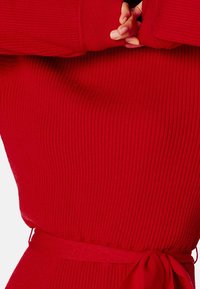 Pull rouge à côtes avec une coupe ajustée, manches longues et taille à lien. La texture du tissu est douce avec de subtiles lignes horizontales.