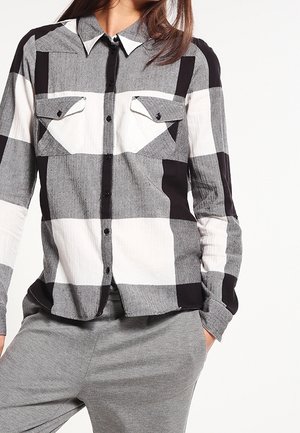 Kobieta ubrana w czarną, białą i szarą koszulę w kratę zapinaną na guziki z dwoma kieszeniami na piersi, w połączeniu z szarymi spodniami, ręce w kieszeniach.