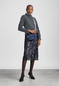 Mulher vestindo um suéter cinza de gola alta, saia midi navy com lantejoulas, meias pretas, sapatos de salto e uma bolsa transversal de denim com alça estampada contra um fundo liso.