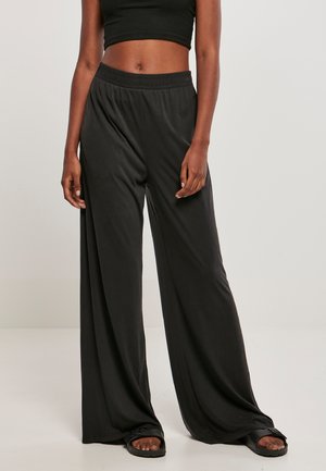 Femme portant un pantalon large noir ample avec une ceinture élastique, un crop top noir et des sandales noires ouvertes à l’avant, debout sur un sol blanc.