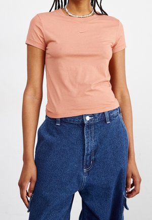 Kvinde iført tætsiddende ferskenfarvet Nike-t-shirt med korte ærmer og lille logo samt højtaljede, løstsiddende blå cowboybukser, stående med afslappede arme.