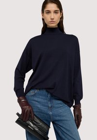 Maglione a collo alto navy con spalle abbassate, abbinato a jeans blu e guanti in pelle bordeaux che tengono una pochette nera texturizzata.