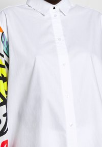 Witte blouse met een puntkraag en kleurrijke patroonmouwen met gedurfde ontwerpen in geel, roze en blauw.