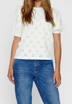 Mujer vistiendo una blusa blanca de manga corta con patrones cortados en forma de cuadrados combinada con jeans de mezclilla azul, mostrada desde el cuello hasta las caderas.