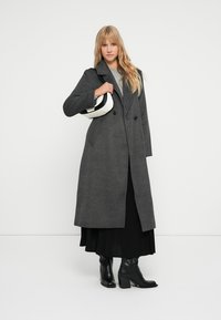 Abrigo de lana gris con cierre de doble botonadura, amplios solapas, bolsillos frontales y una textura suave, combinado con una falda plisada negra y botas negras.