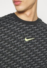 Černé tričko s opakujícími se šedými logy Nike, kulatým výstřihem a krátkými rukávy. Na hrudi se nachází jasně žluté logo Nike.