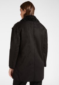 Donna vista da dietro mentre indossa un cappotto in finta pelliccia nera con dettagli delle cuciture visibili e colletto testurizzato su uno sfondo bianco puro.