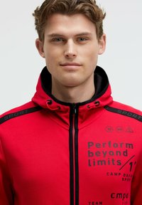 Sudadera roja con acentos negros, cierre completo, cuello alto y gráficos impresos que dicen "Rinde más allá de los límites". Hecha de un tejido suave.