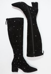 Mustat mokkasaappaat polveen saakka, joissa on matalat lohkokengät, koristeelliset hopeiset niitit ja sivuvetoketjut valkoisella taustalla.