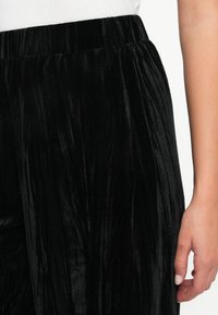 Vrouw draagt zwarte geplooide fluwelen broek met elastische tailleband en witte top, hand ontspannen naast haar lichaam.